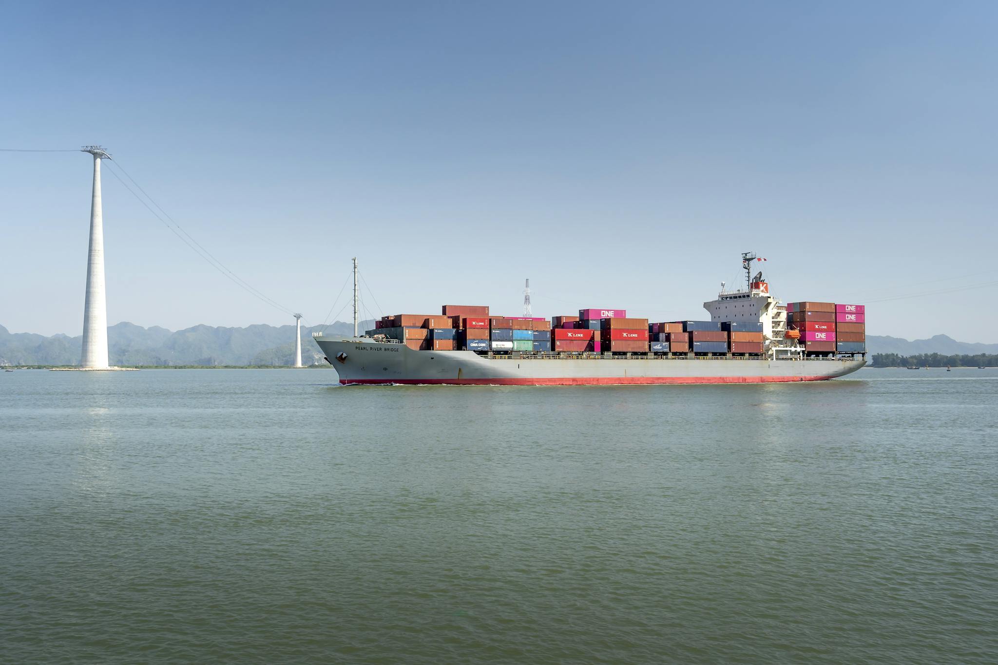 A large cargo ship carrying colorful containers sails smoothly across a calm ocean on a clear sunny day.