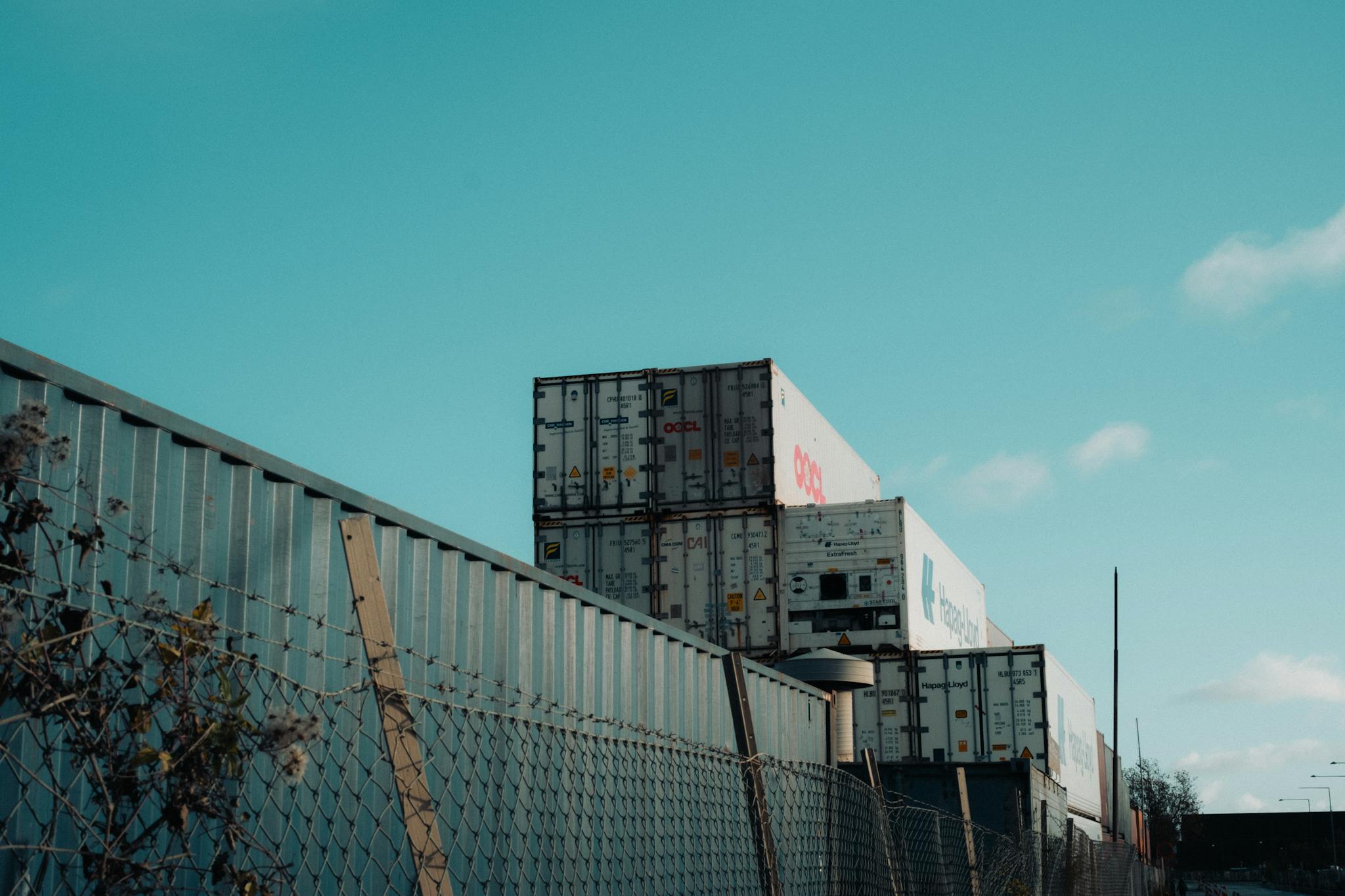 A view of stacked shipping containers beside a fence under a clear blue sky, captured outdoors.